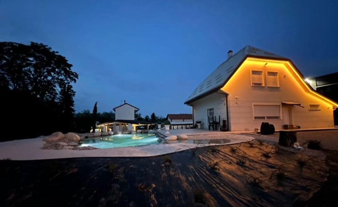 Casa e piscina illuminate al crepuscolo, accanto a un patio buio e agli alberi.