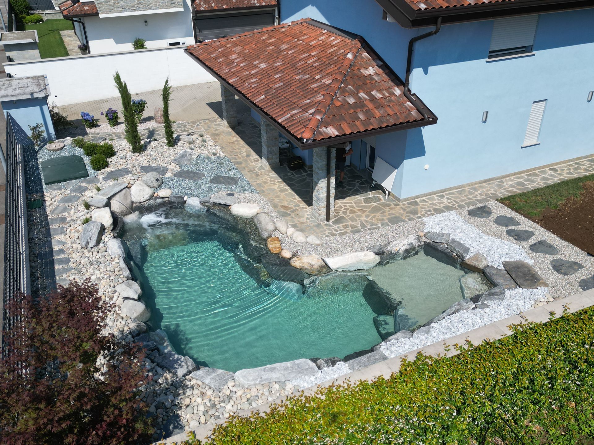 Piccola piscina nel cortile sul retro, accanto a una casa blu, con patio in pietra e tettoia con tegole rosse.