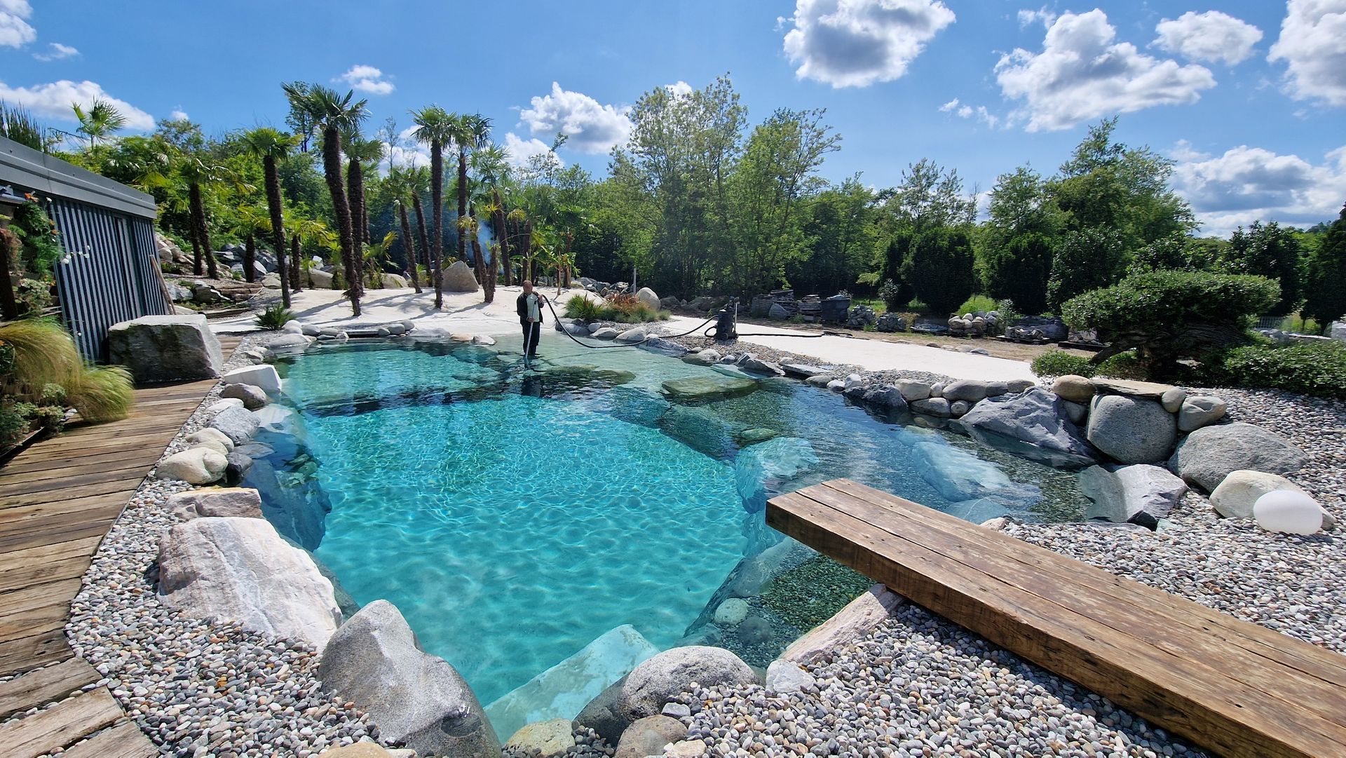 Piscina naturale all'aperto con acqua turchese, rocce, alberi e una passerella di legno sotto un cielo soleggiato