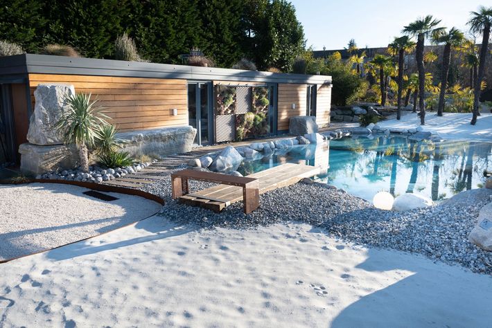 Piscina moderna del resort con pavimentazione in pietra, lettini e palme accanto a un edificio rivestito in legno.