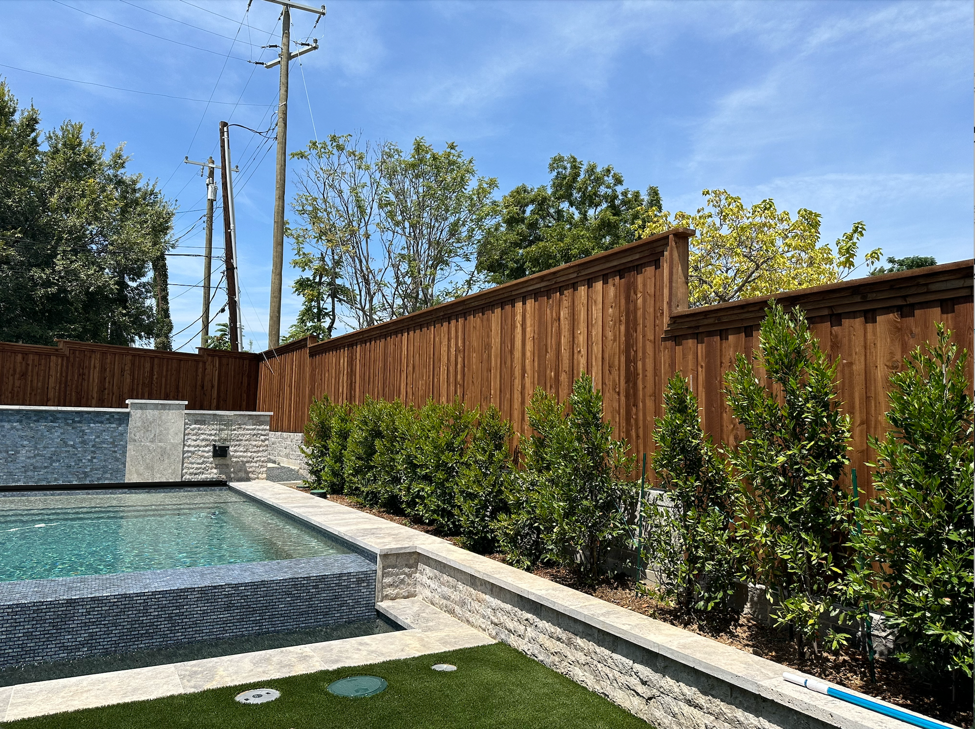Backyard pool beside a wooden fence, landscaped shrubs, and blue sky
