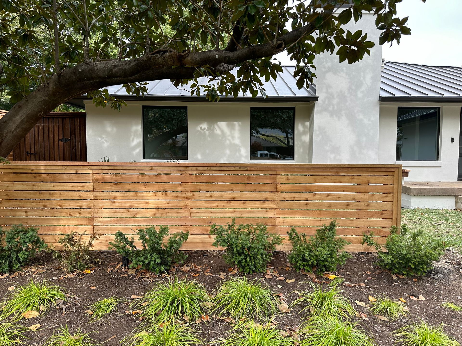 Horizontal wooden fence with greenery, in front of a white house with dark window frames and a black roof.