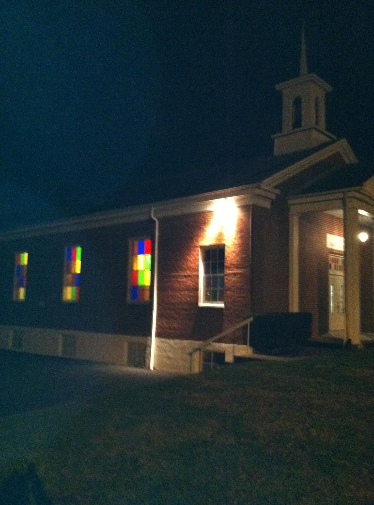 Brick church with steeple, lit windows showing stained glass at night.