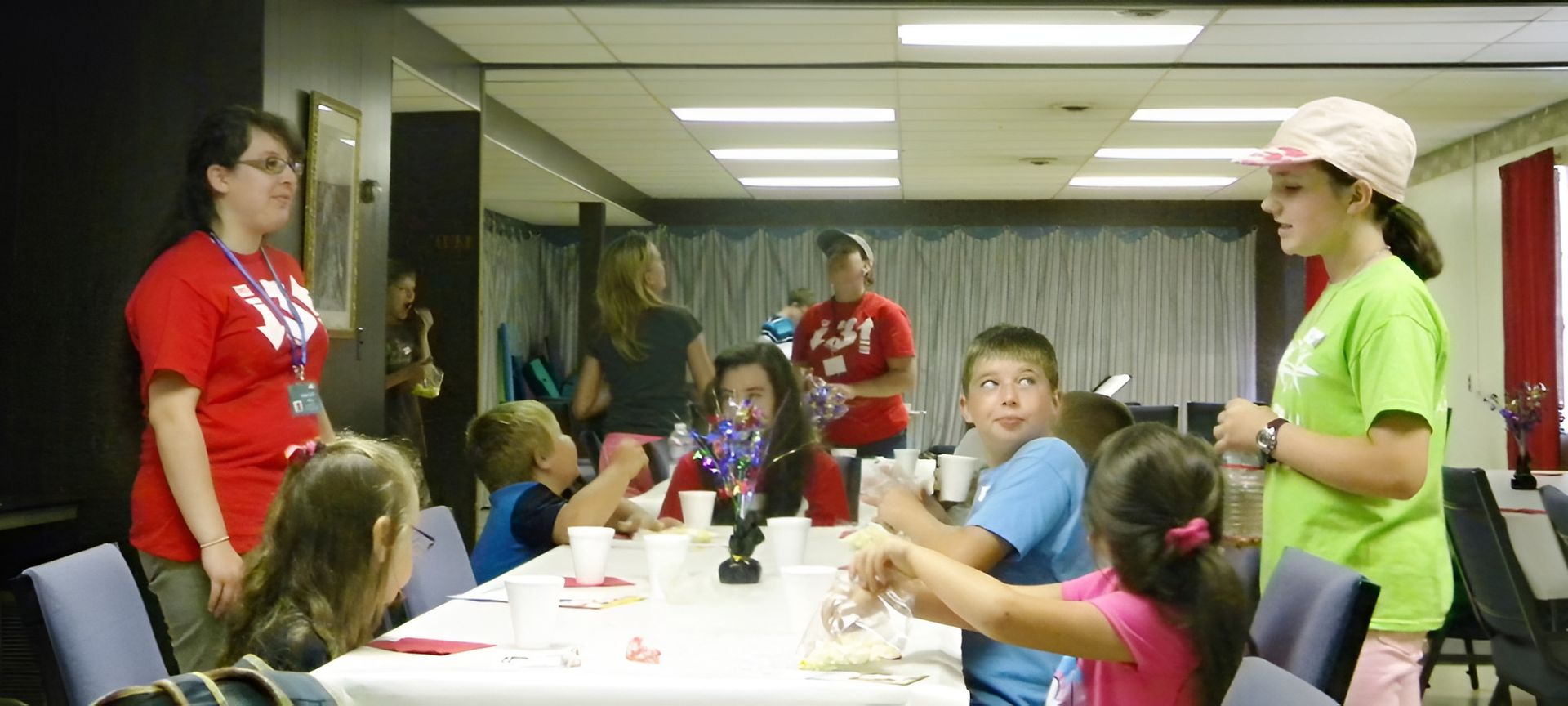 Children and adults at a table in a room. Some wear red or green shirts. A person in a pink hat speaks to the children.