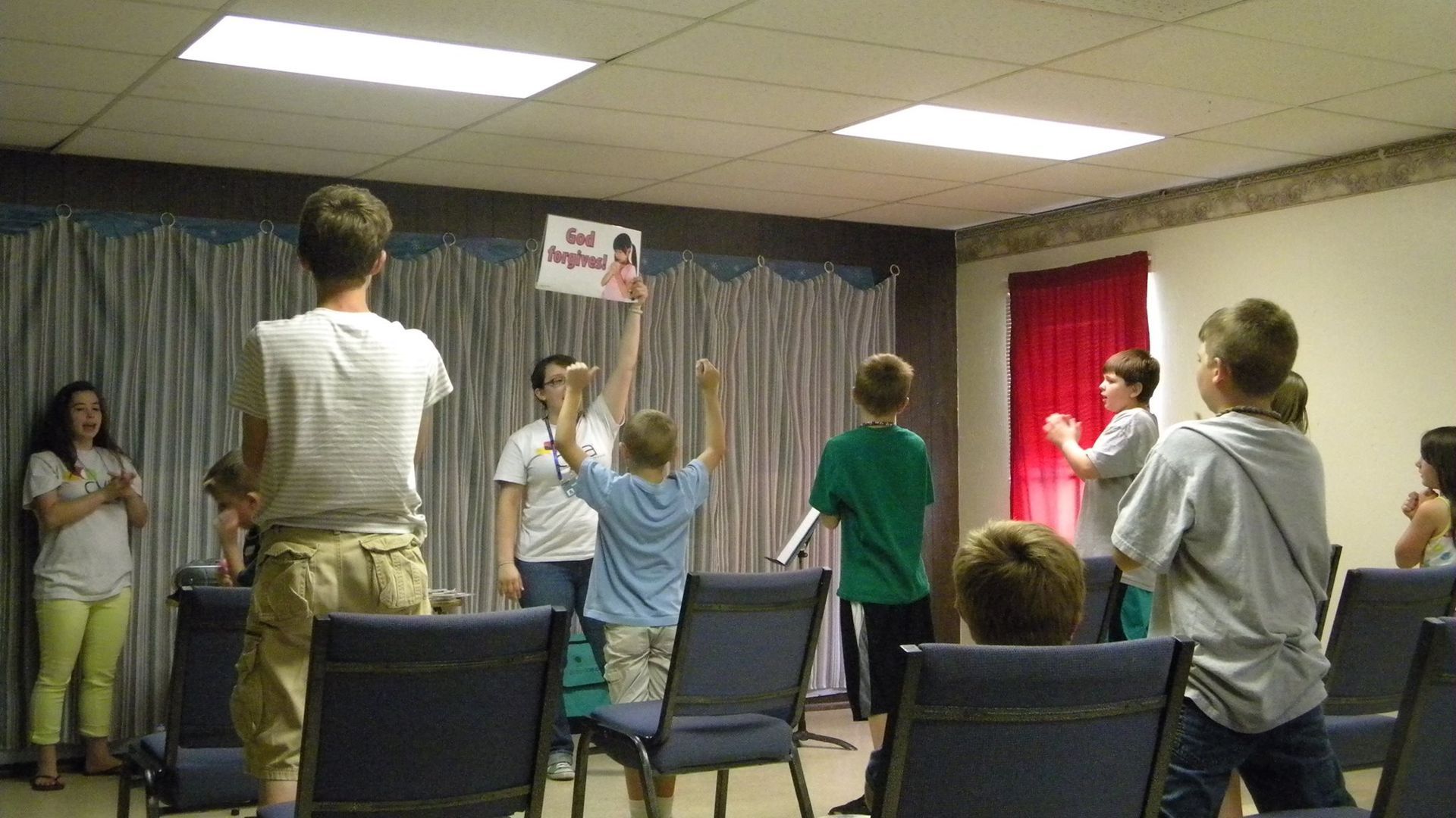Children in room hold up sign, cheer. Others clap. Some stand. Blue chairs in rows.