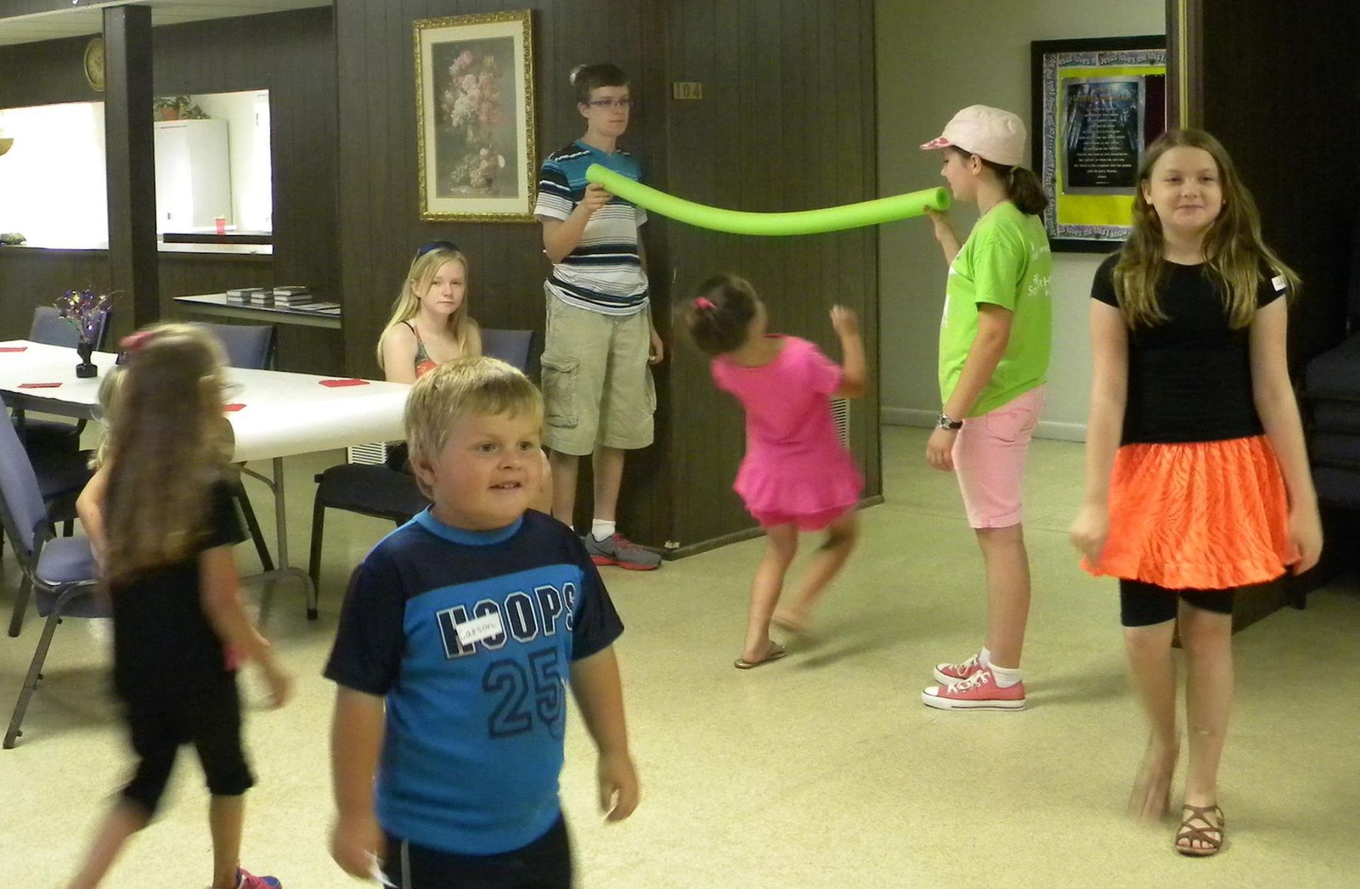 Children playing with a green balloon in a room, some watching, some bending over, and one blowing.