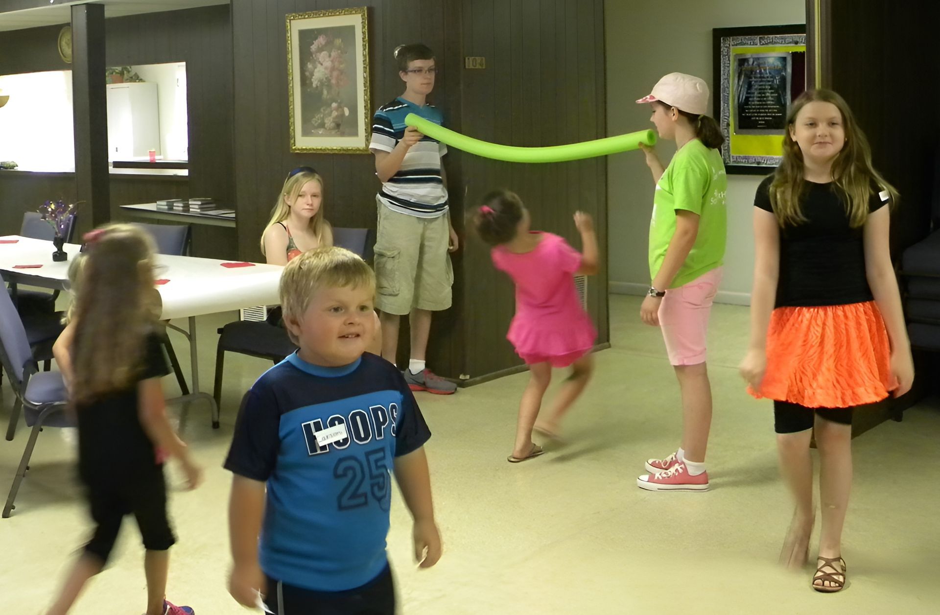 Children playing with a green balloon, in a room with a table, and two people are blowing on it.
