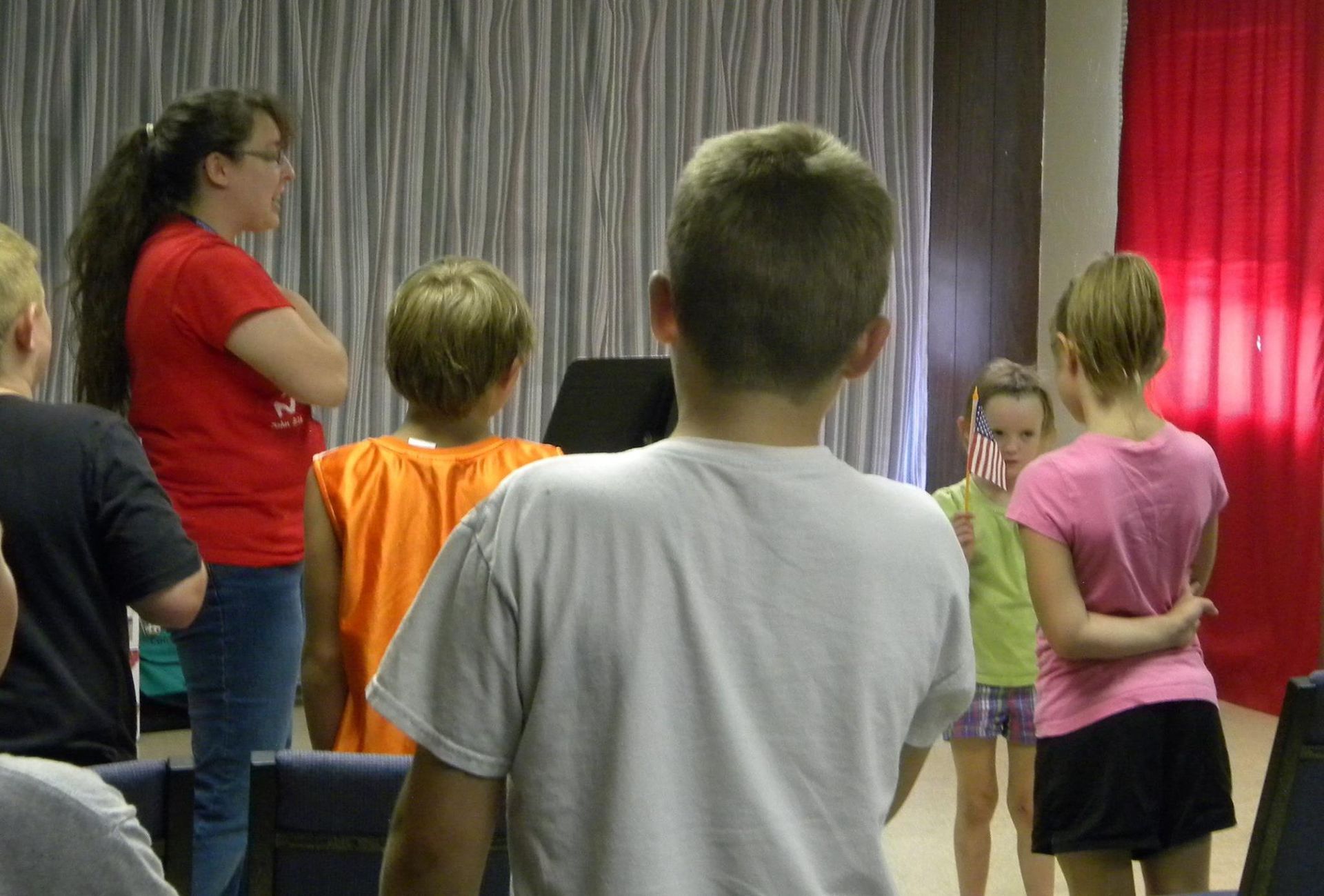Group of children and a woman in a room; some are looking forward, one boy's back is to the camera.