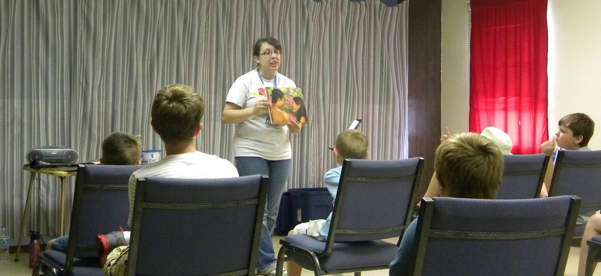 Woman presenting to a group of children, holding a book, standing in front of a striped curtain.