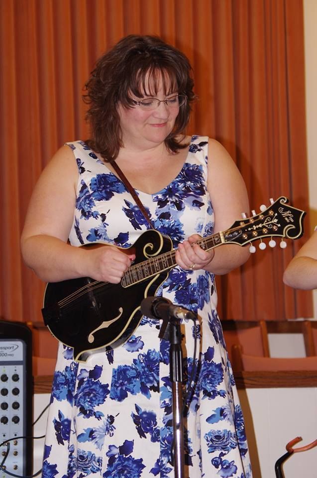 Woman in floral dress playing a black mandolin, standing near a microphone in a room with a beige wall.