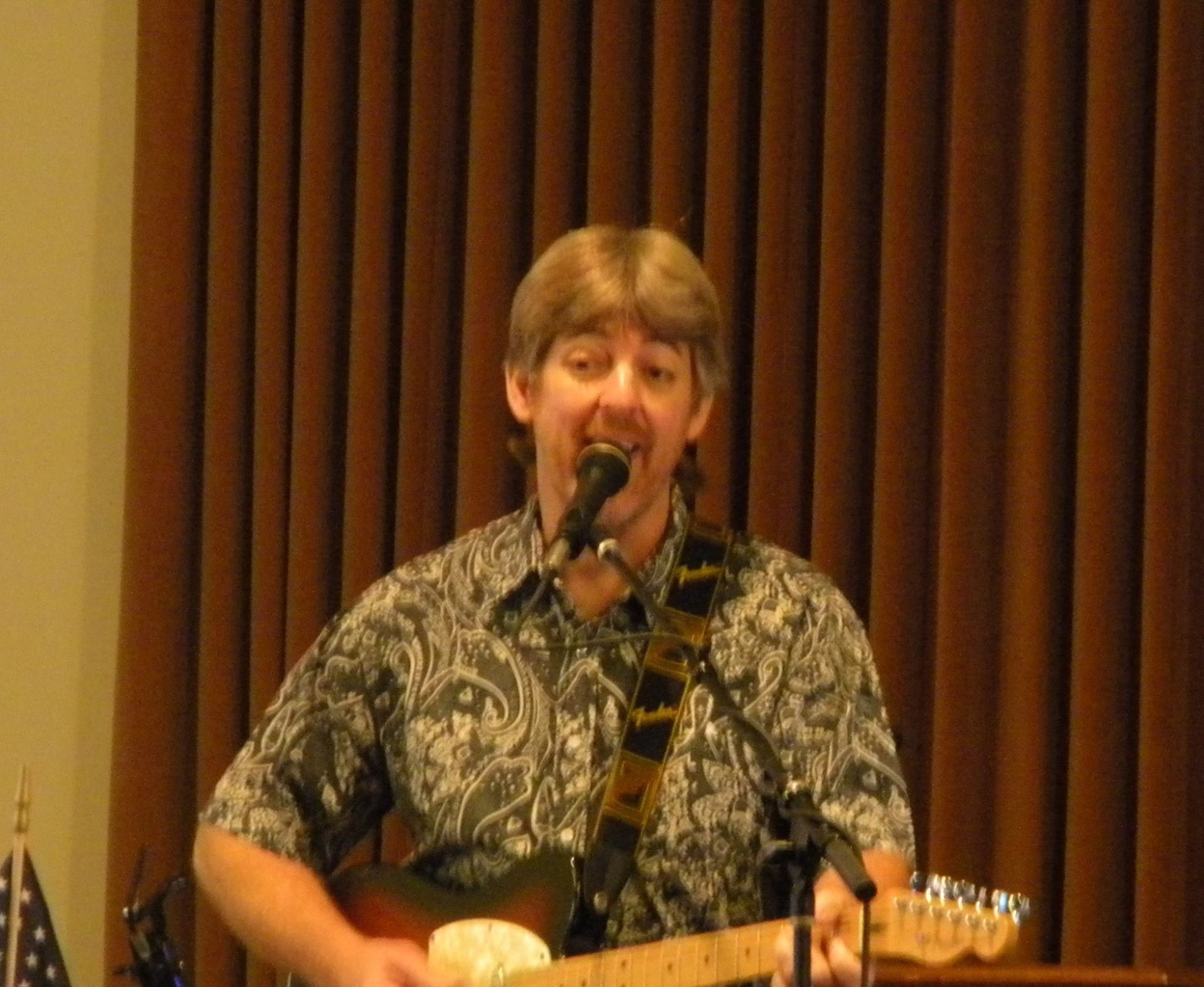 Man singing and playing guitar on stage with brown curtains in background.