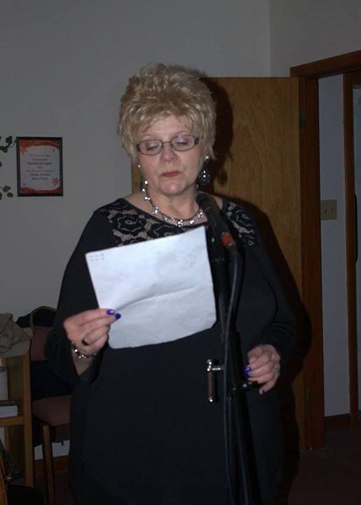 Woman with blonde hair wearing glasses and a black dress reads from paper at a microphone.