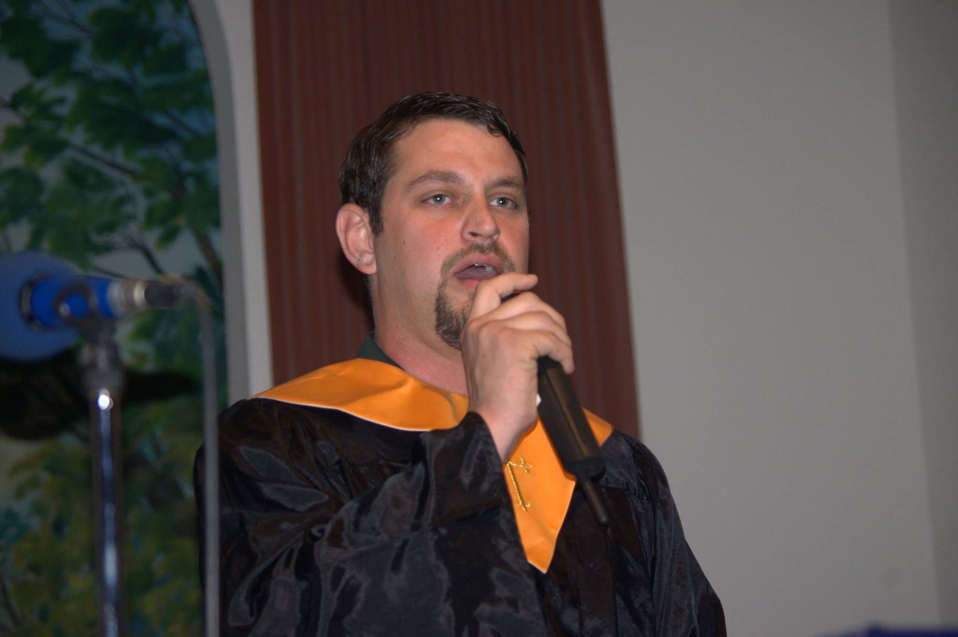 Man in academic robe speaks into a microphone. Dark robe with yellow trim, indoors.