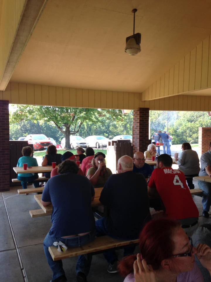 People seated at picnic tables under a covered shelter, with cars and trees visible in the background.