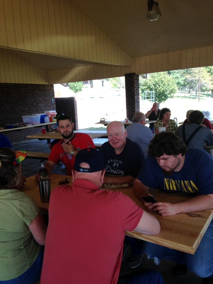 People sit at a picnic table under a pavilion. Several are looking at phones, others are talking. Outdoors on a sunny day.