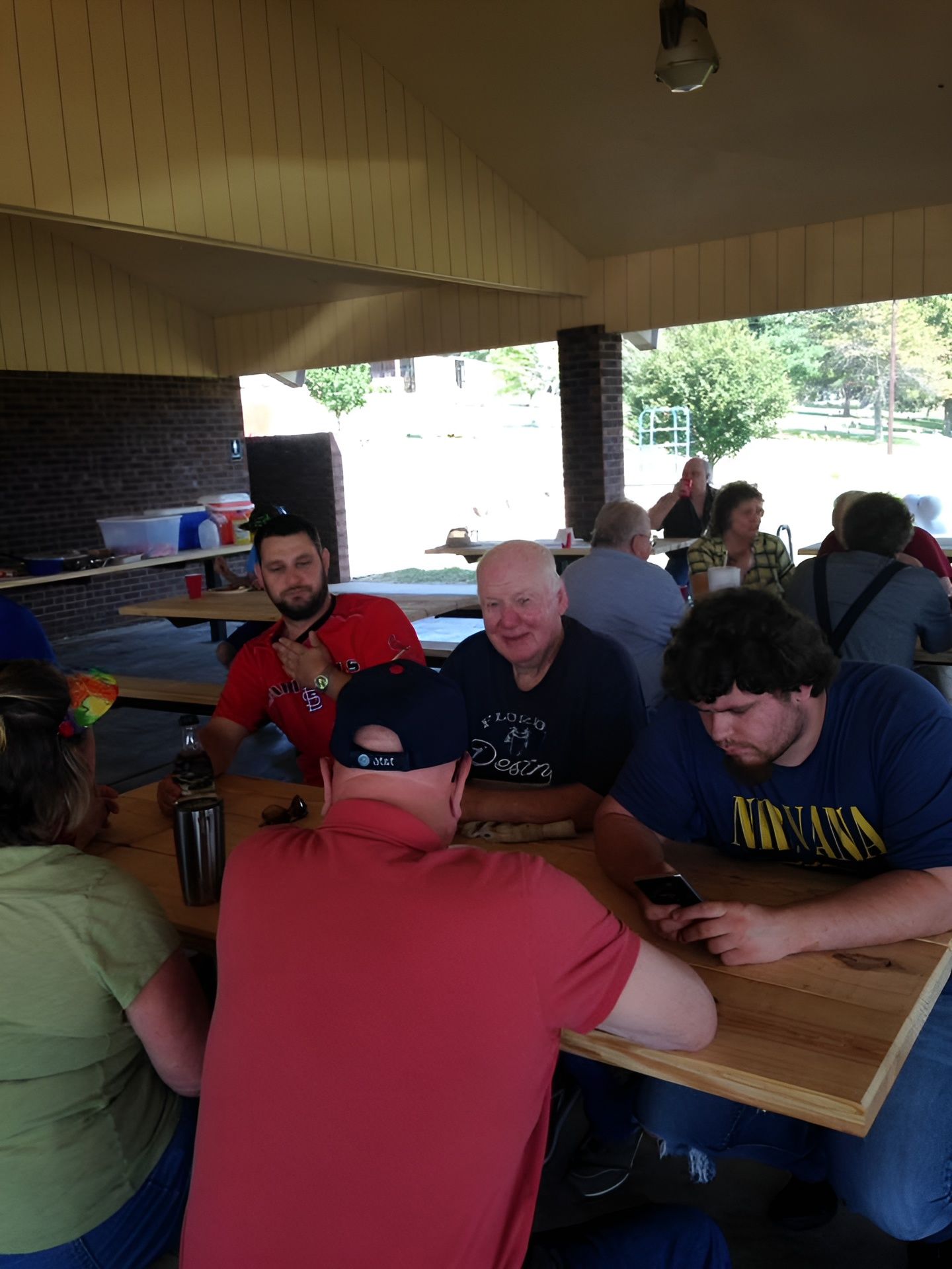 People seated around a picnic table under a pavilion. One person looks at their phone; others converse.