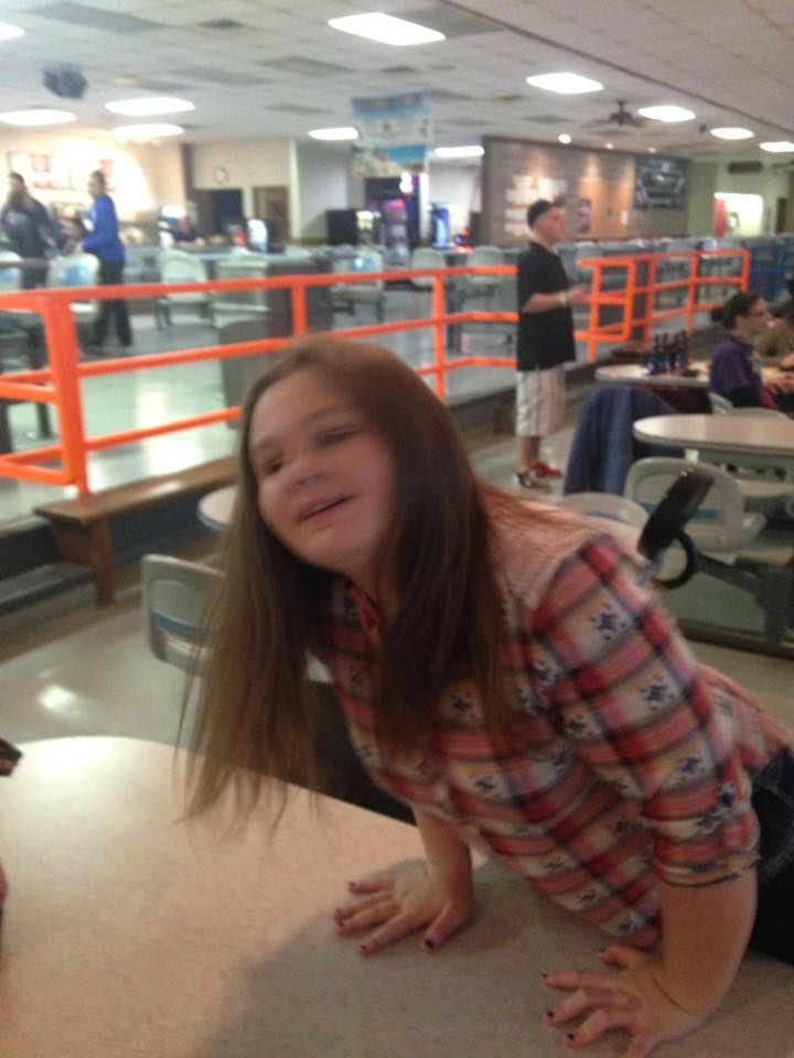 Young person leaning on a bowling alley table, smiling. Wearing a plaid shirt, bowling alley in background.