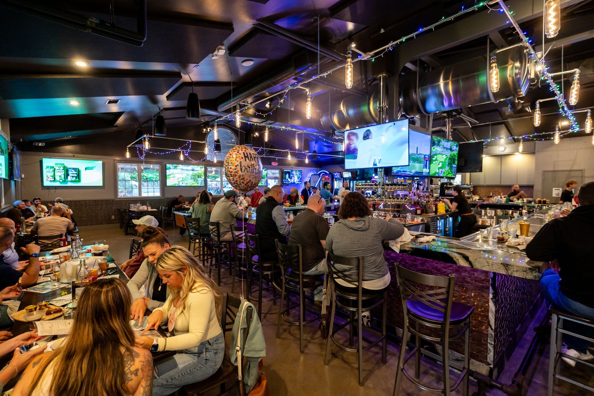 Busy bar and restaurant interior with patrons dining, glowing TVs, and blue-purple neon lighting.