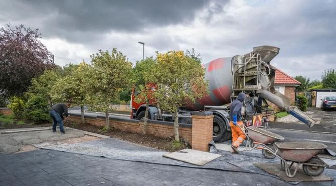 Workers pour concrete beside a cement mixer at a residential construction site with autumn trees.