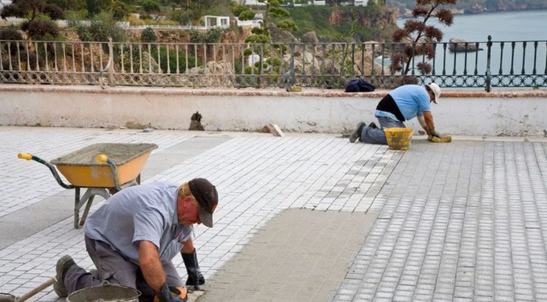 Two workers laying gray paving tiles on a seaside walkway, with wheelbarrows nearby.