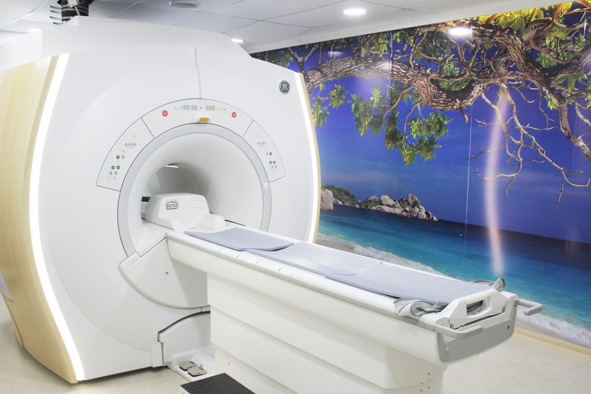 Aparelho de ressonância magnética em ambiente médico com uma maca, de frente para uma parede com um mural de praia.