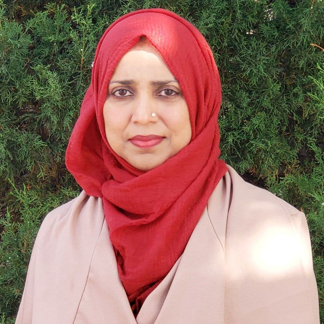 A woman wearing a red hijab and a tan jacket is standing in front of a bush.