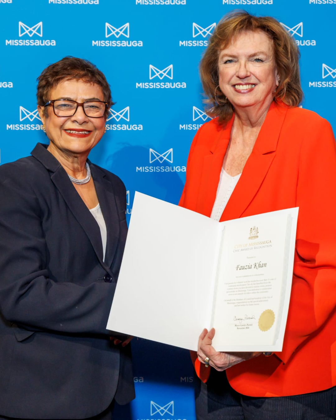 Two women in front of a blue backdrop, one presenting a framed award. One wears a blue suit, the other a red blazer.