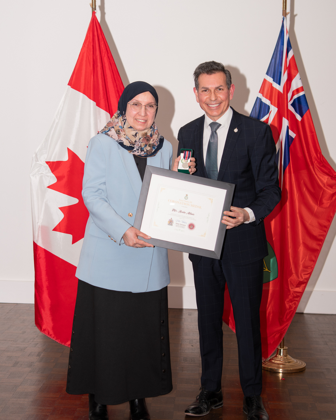Woman in headscarf and blue coat receives framed award from man in suit, with Canadian and Ontario flags behind them.