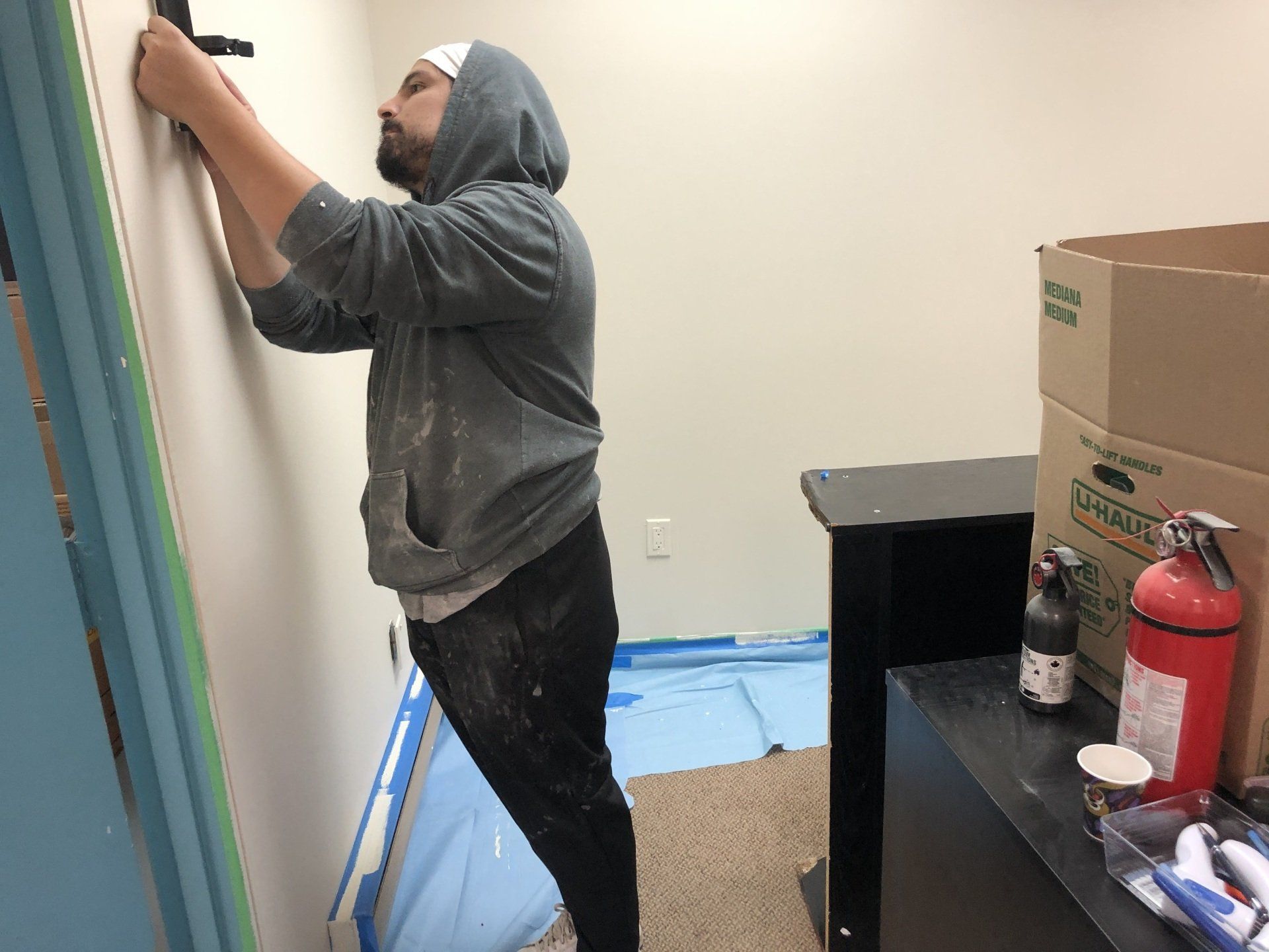 A man is painting a wall in a room with boxes and a fire extinguisher.