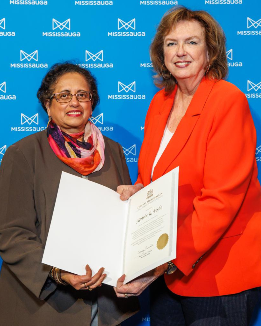 Two women in front of a blue backdrop, one presenting a certificate. One wears an orange jacket, the other a brown coat.