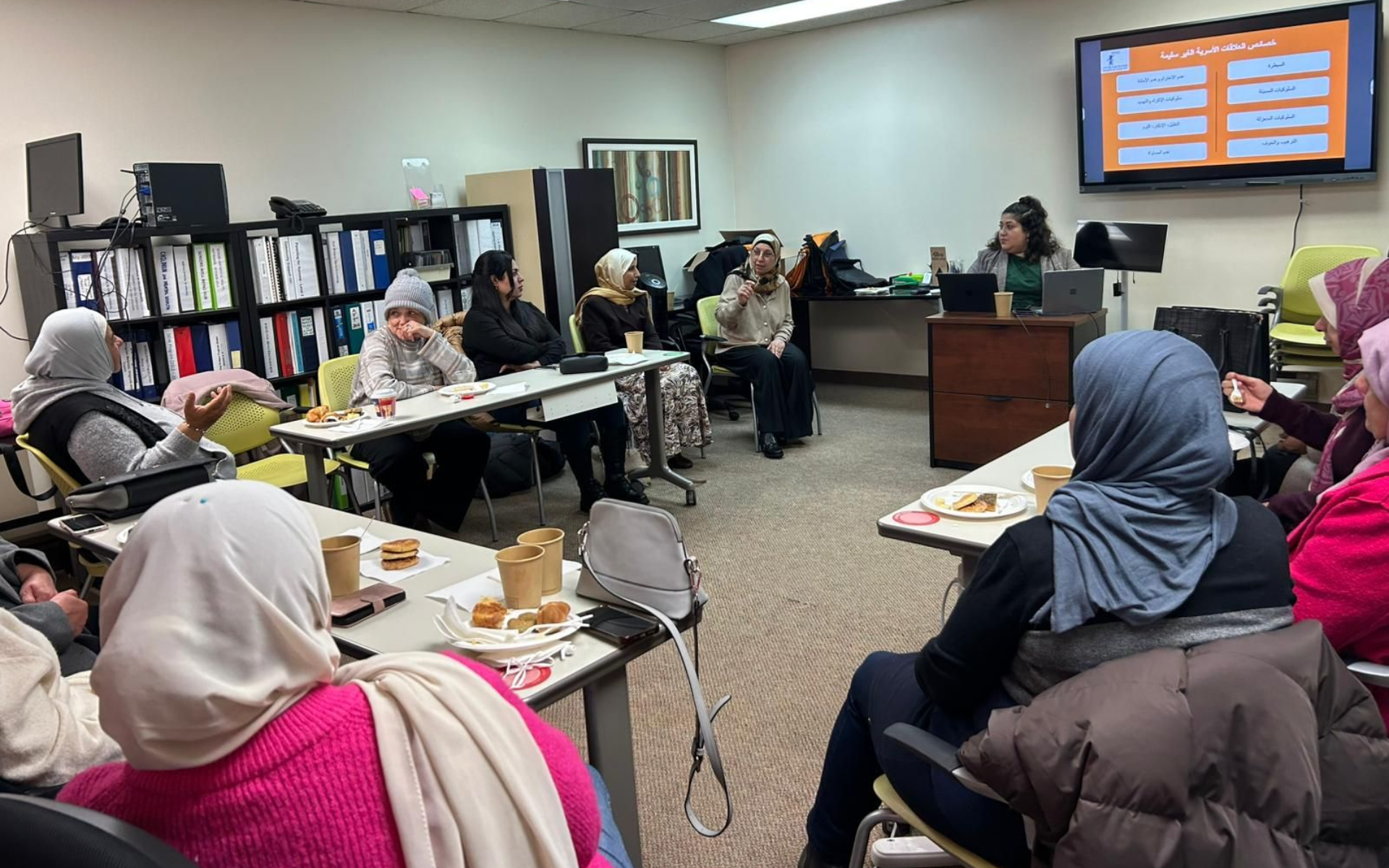 Group of people in a meeting. A woman is presenting at the front with a screen displaying text. Tables with food items.