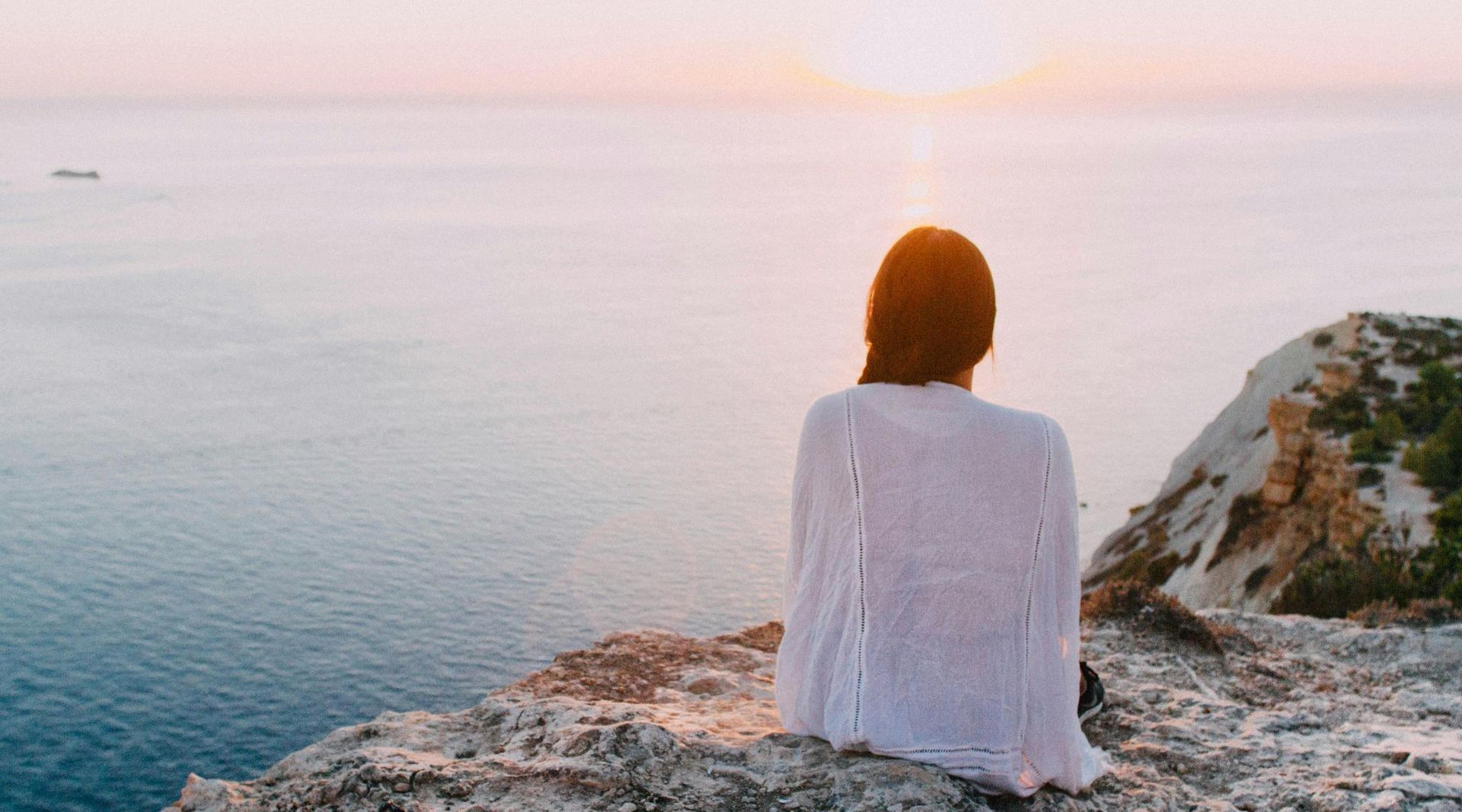 Woman in white sits on cliff, looking out at ocean and setting sun.