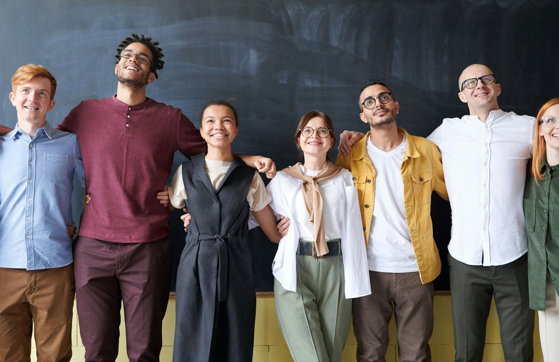 Group of diverse people with arms around each other, smiling, in front of a chalkboard.