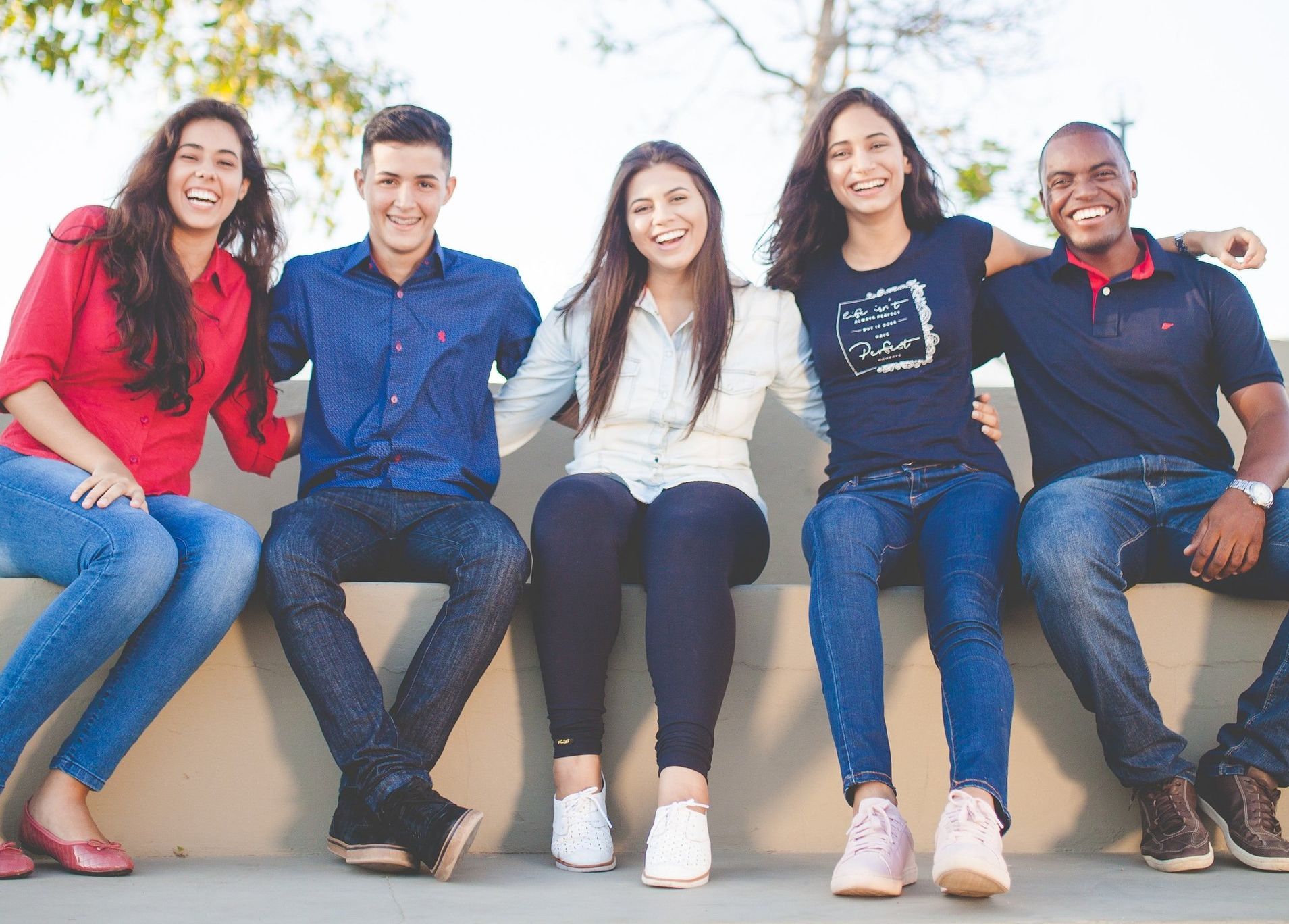 Five diverse young adults smiling, sitting closely together outside, arms around each other.