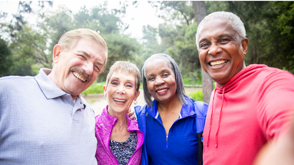 Four smiling older adults in a park. One takes a selfie, others have arms around each other.