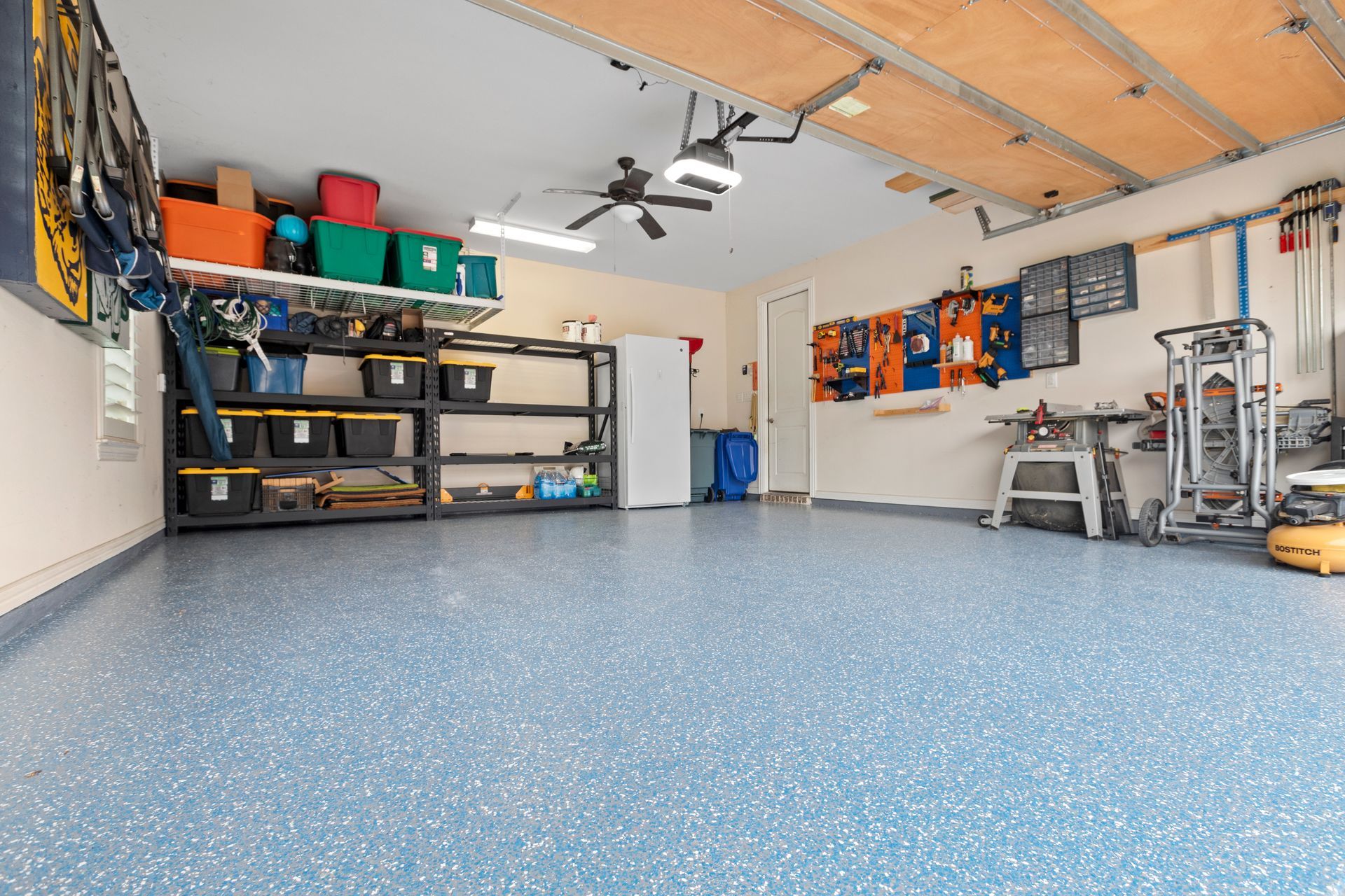 Spacious garage with speckled blue floor, storage shelves, workbench, and tools under an open ceiling.
