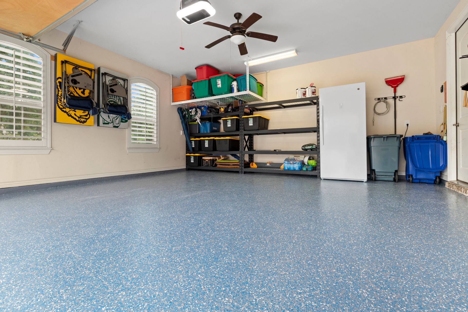 Empty garage with speckled epoxy floor, wall shelves, white cabinet, windows, and a ceiling fan
