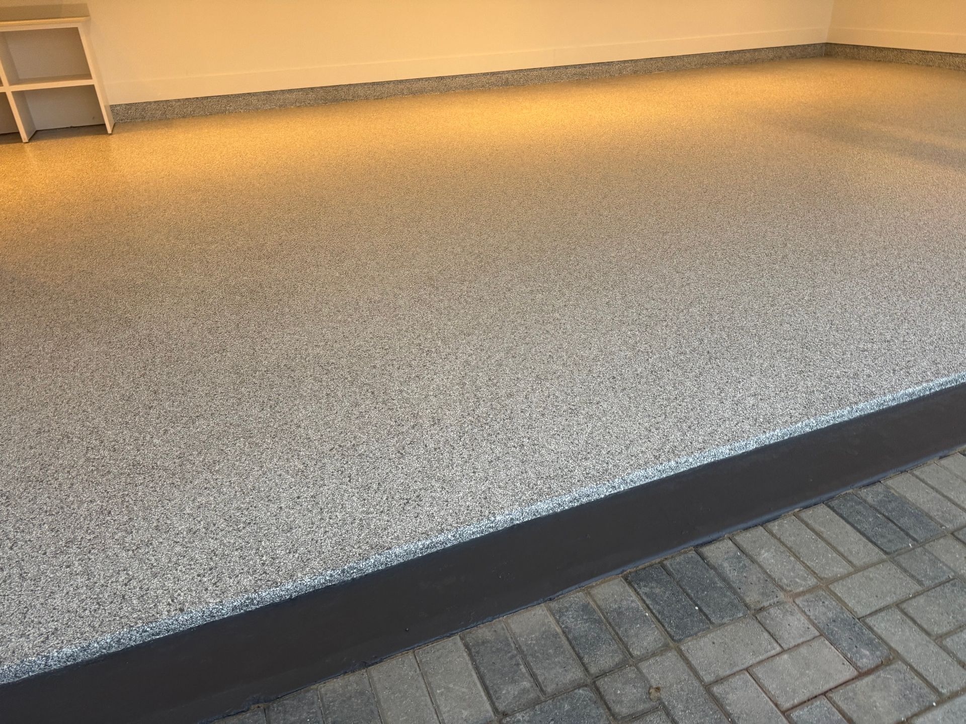 Empty raised pebble-textured patio beside a brick paver walkway, lit by warm indoor light