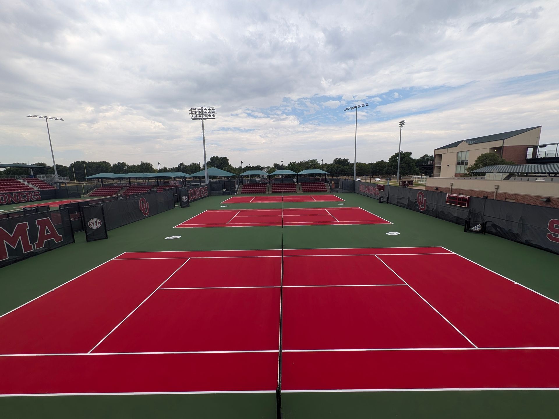 Empty red tennis courts under a cloudy sky, viewed from behind the net.