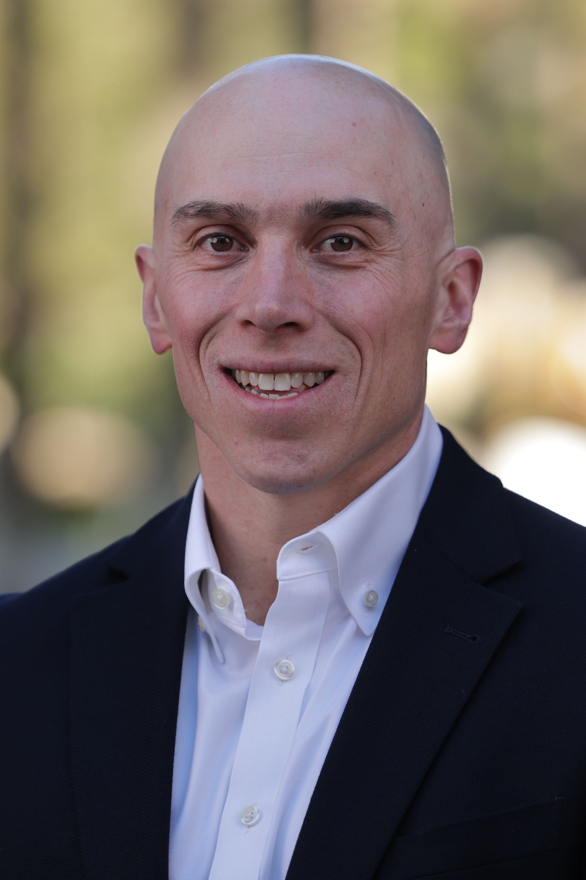 Smiling bald man in a dark blazer and white shirt, standing outdoors with a blurred background