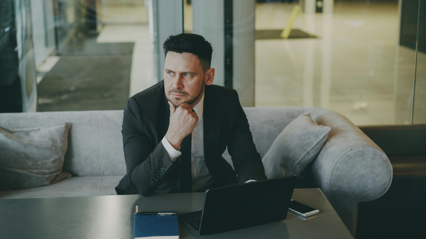 Man in a dark suit sitting at a table with a laptop, resting his chin on his hand in a modern office setting