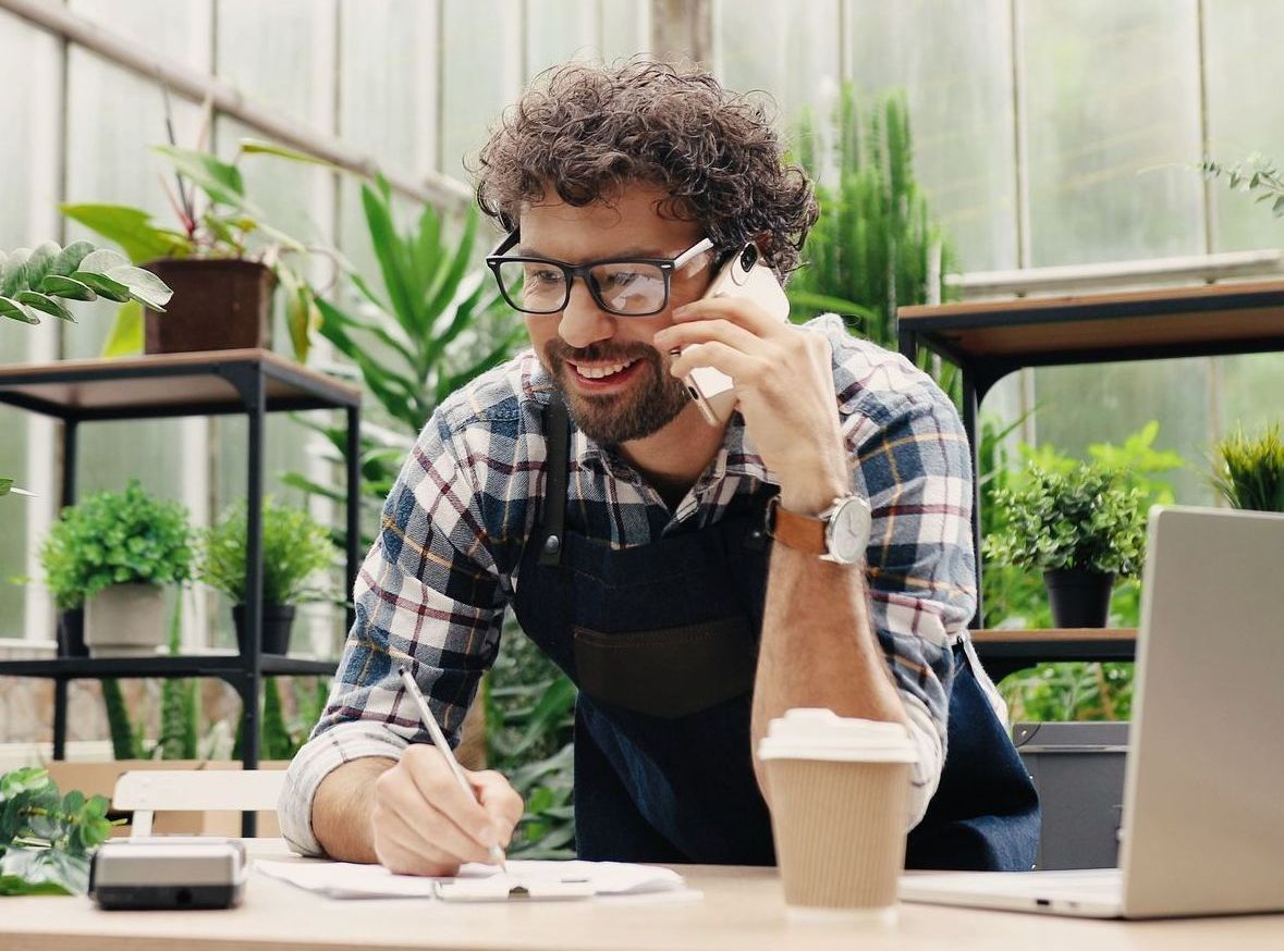 Person talking on a phone at a table in a greenhouse with plants and a laptop nearby