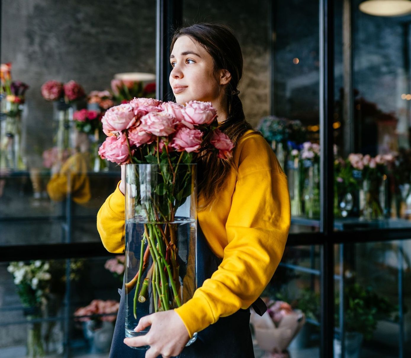 Woman in a yellow sweater holding a large bouquet of pink flowers in a glass vase at a flower shop