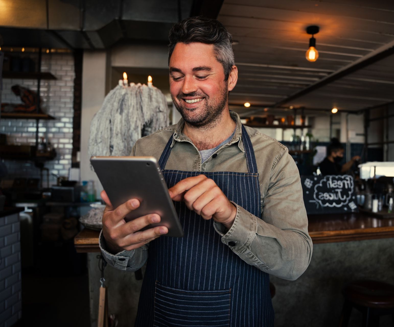 Smiling café worker in an apron using a tablet behind the counter