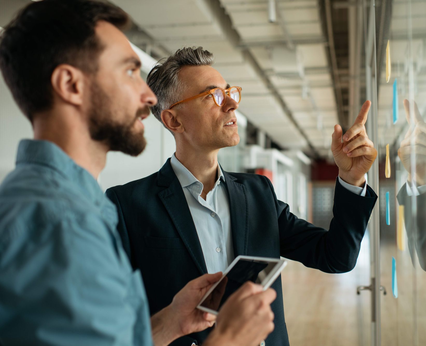 Two colleagues review notes on a hallway wall, one pointing while holding a tablet.