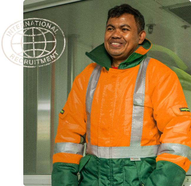 A man wearing an orange jacket and green gloves stands in front of a sign that says international recruitment