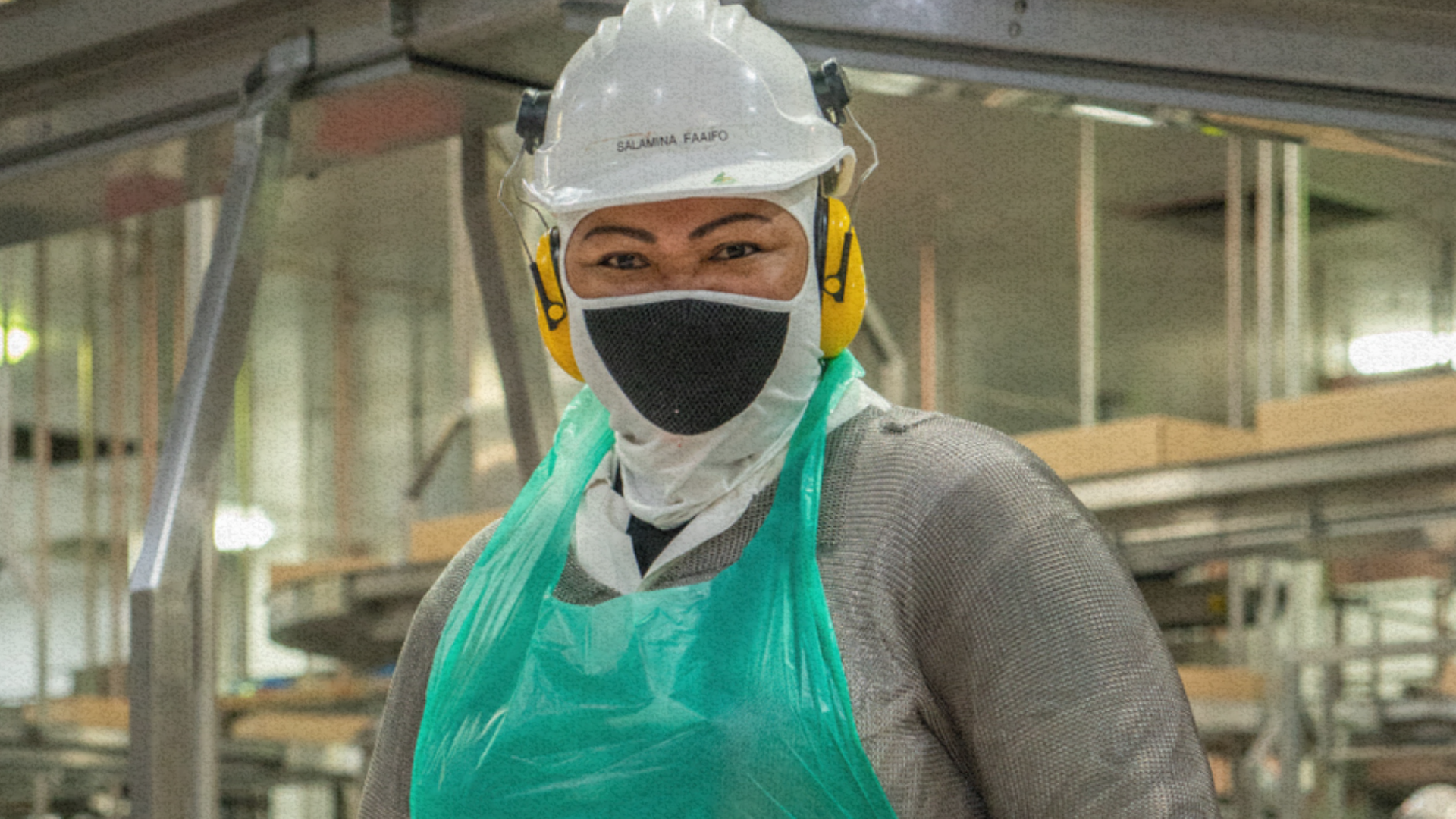 Smiling worker in protective gear, including a hard hat, face mask and apron, standing in an industr