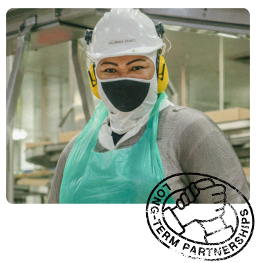 Smiling worker in protective gear, including a hard hat, face mask and apron, standing in an industrial setting. 'Long-Term Partnerships' stamp in the corner.