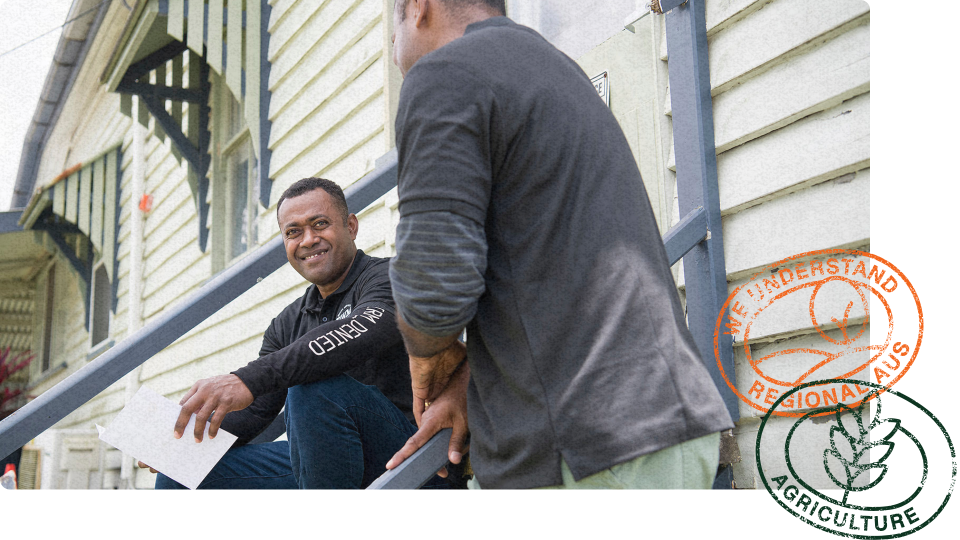Two men in casual workwear sit on outdoor steps, engaged in conversation, with one holding a document and smiling.