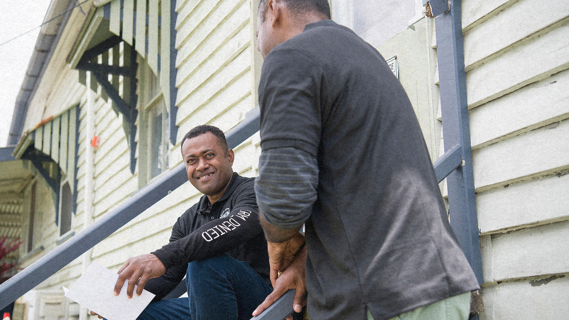 Two men in casual workwear sit on outdoor steps, engaged in conversation.